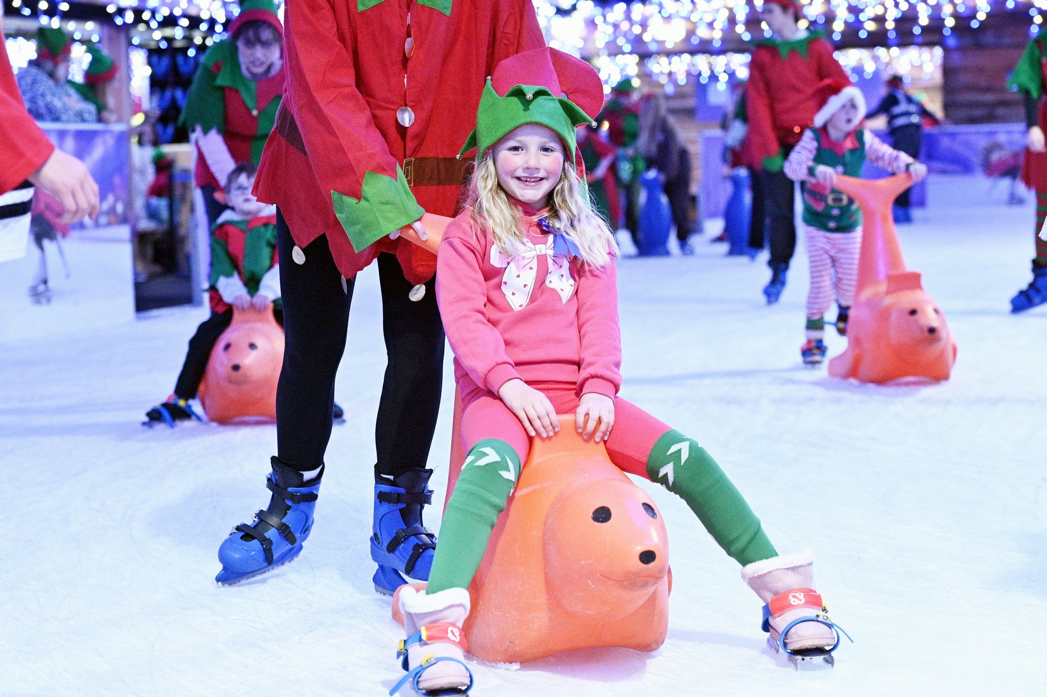 A girl dressed an elf sitting on an orange seal skater aid enjoying ice skating. 
