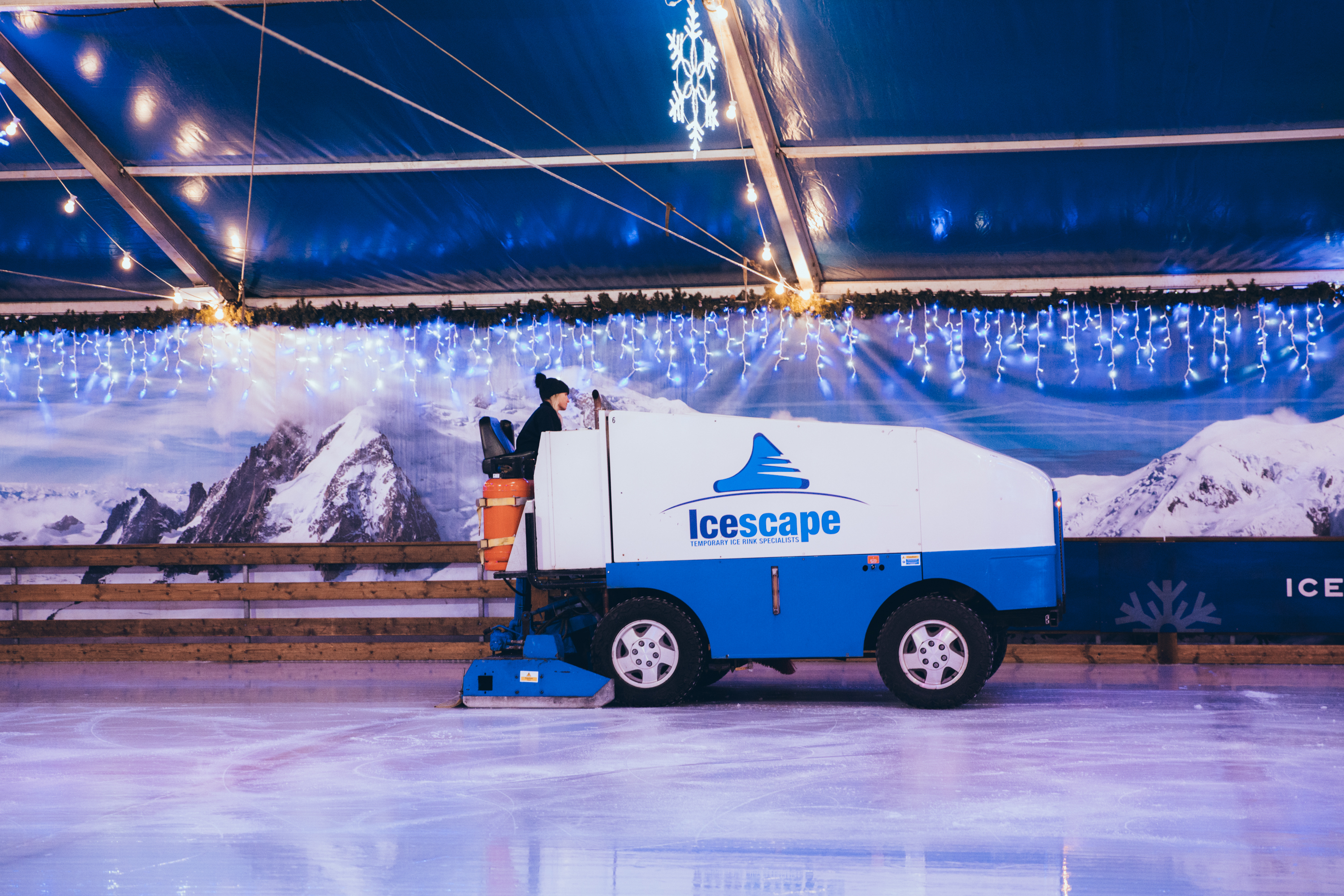 An Icescape Olympia machine at work maintaining the temporary real ice rink. The machine is on the ice with wooden barrier and alpine mountains backdrop in the background. 