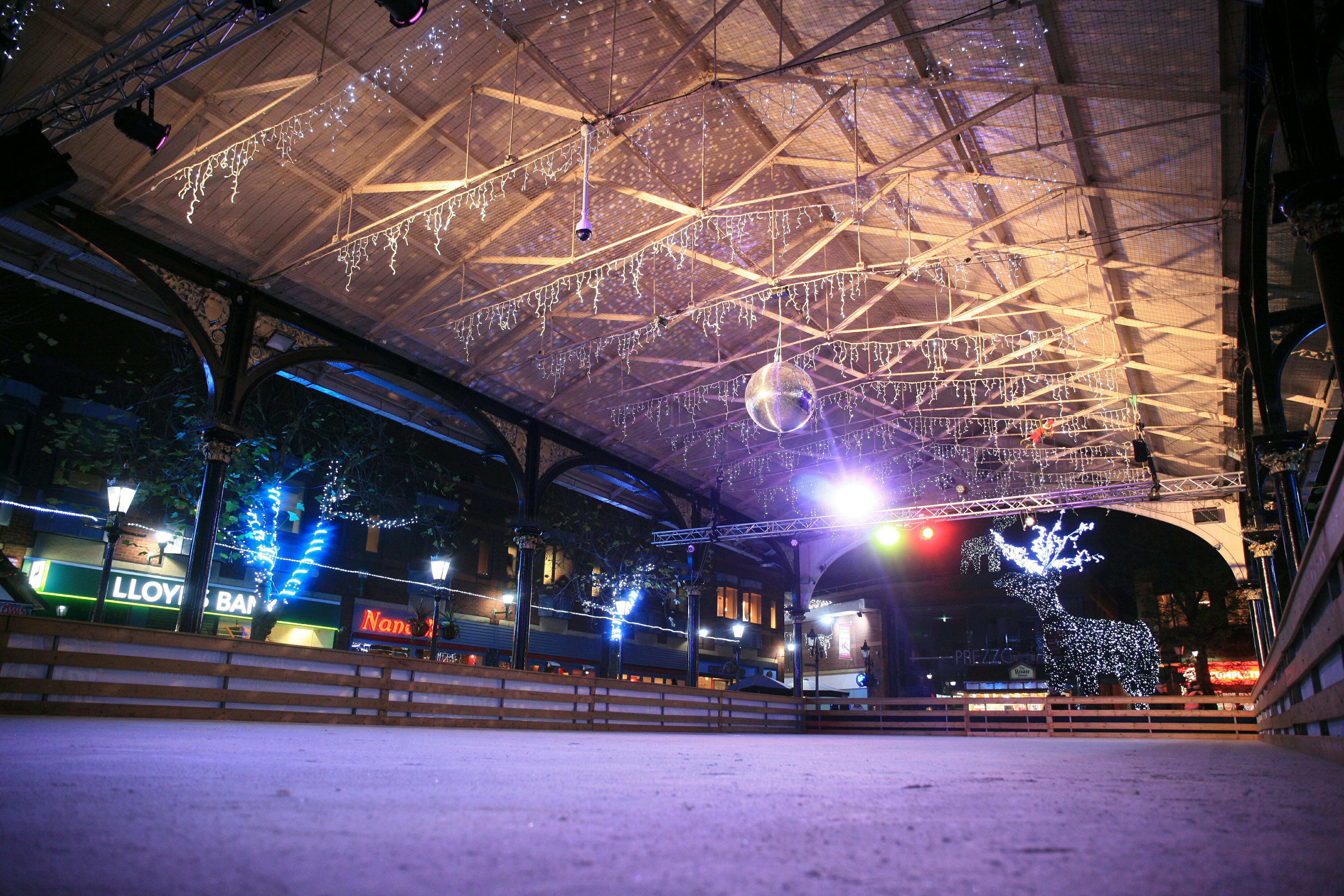 A winter ice rink at night. The ice is empty with lights a disco ball above. A large lit up Reindeer is at the end of the ice rink. 