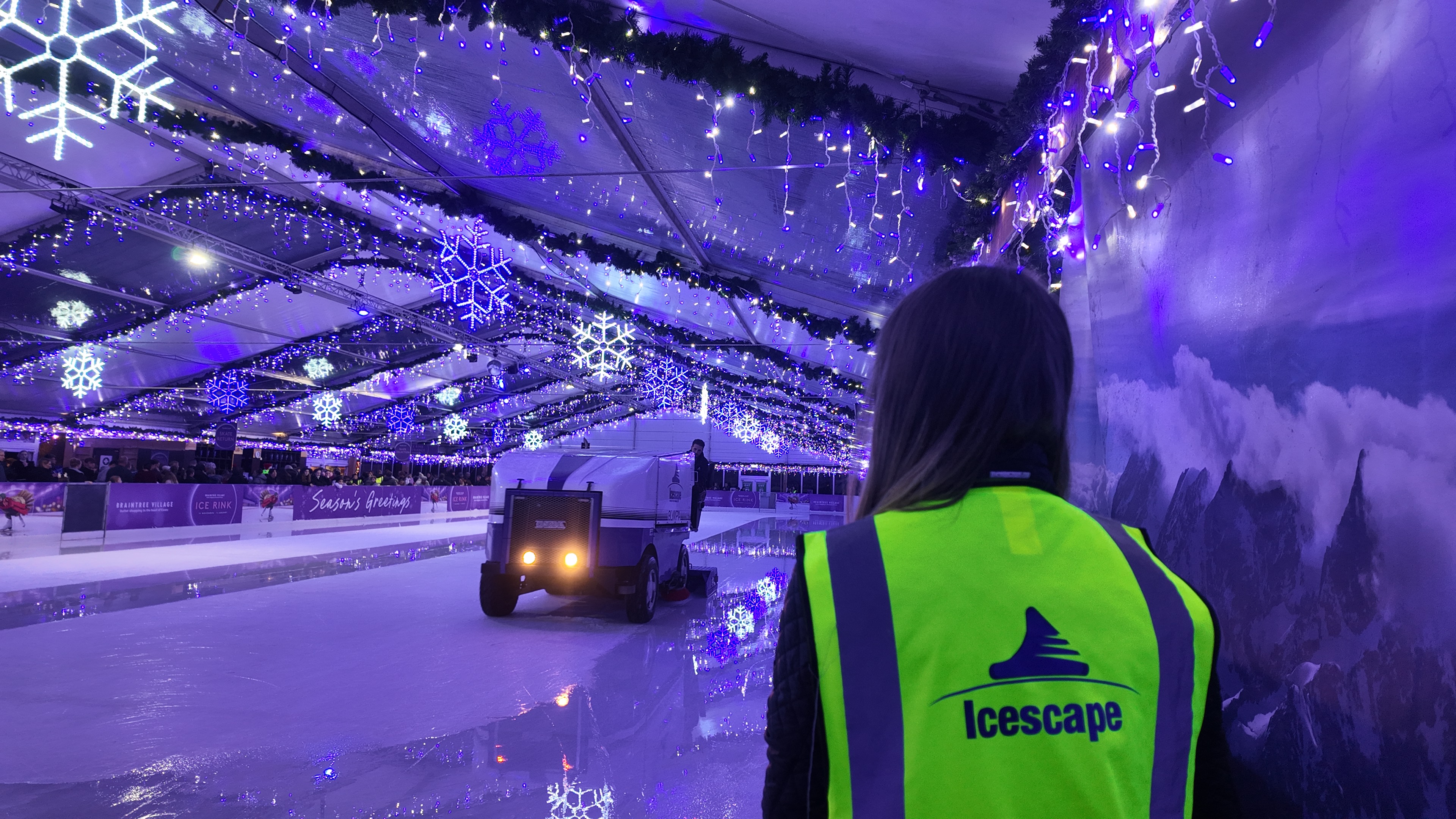 An ice resurface at a winter ice rink. The machine is laying water on the ice rink, whilst a staff member in hi-vis is waiting to clear a corner. 