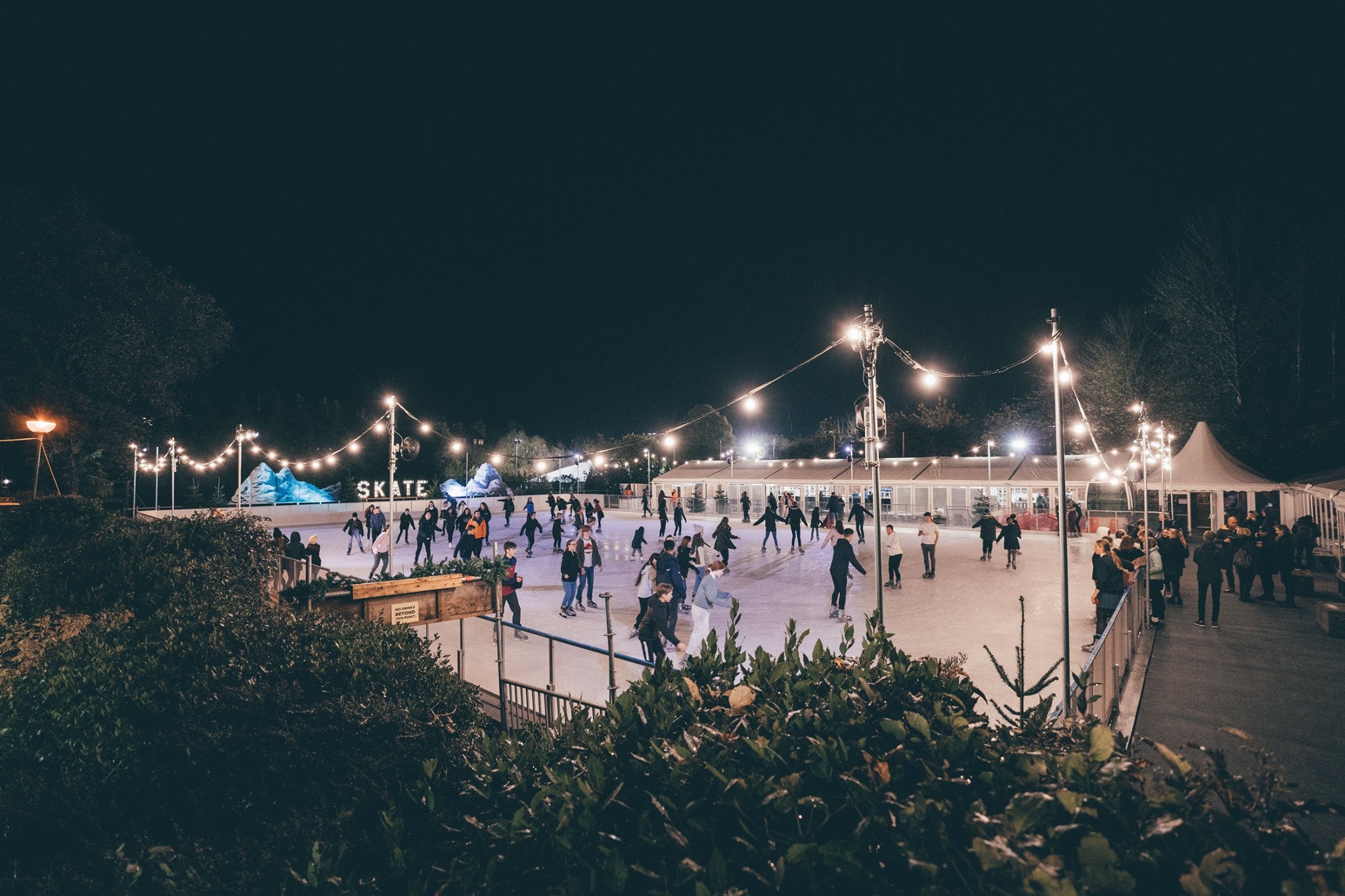 Skaters enjoying skating at Bath on Ice in the night time with bright festoon lights surrounding. 